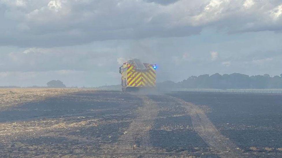 The yellow and red back view of a fire engine, with its tyre tracks crossing a blackened field of stubble. Smoke has greyed out the image as it billows across the field.