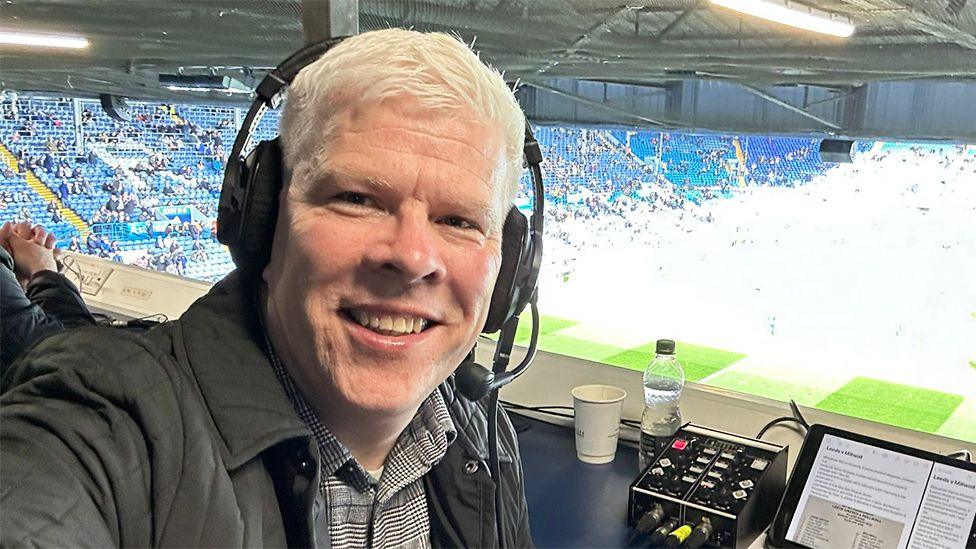 Bryn Law sitting on a gantry, with headphone on. He has a screen on the desk with the green football pitch behind him bathed in sunshine.