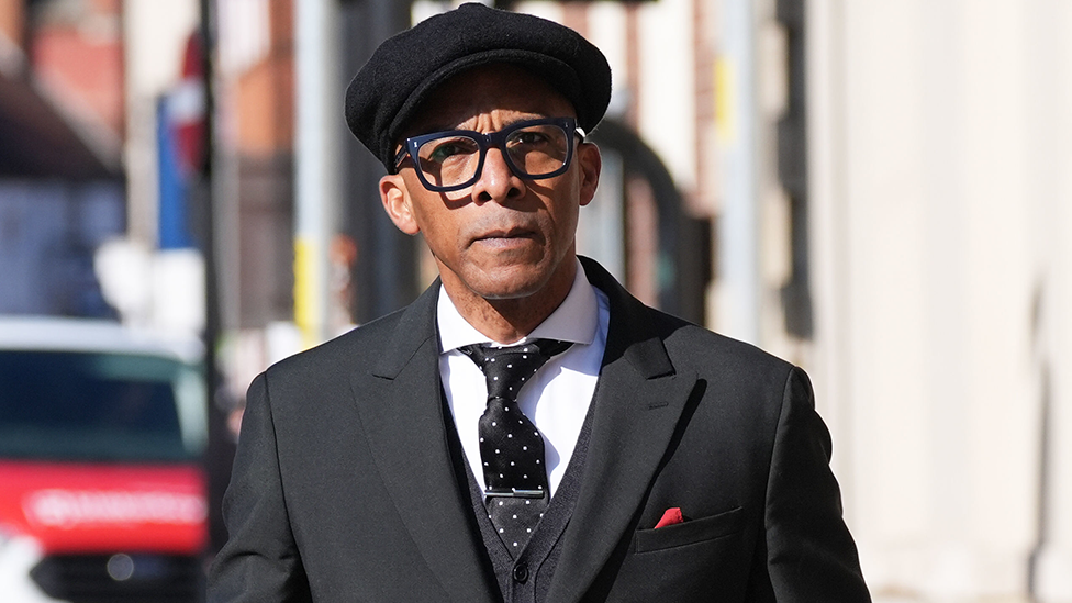 Image shows Jay Blades arriving at Worcester Crown Court wearing a black suit, white shirt, black tie with white spots and a tie pin and a red handkerchief tucked into his breast pocket. He is wearing a black peaked cap.