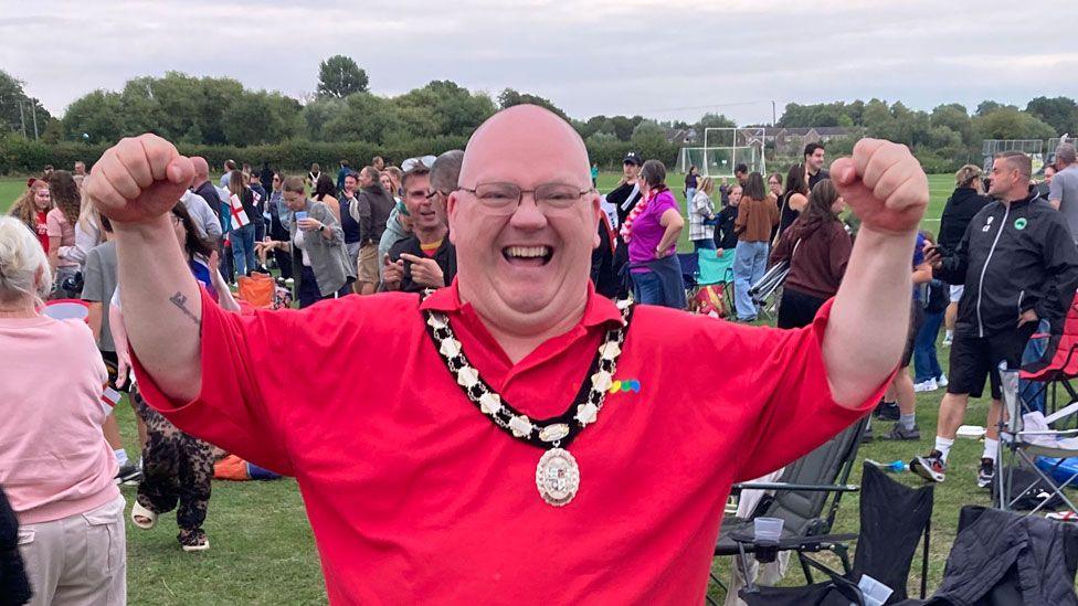 Paul Day standing in a park in Newport Pagnell. He is wearing a red England shirt, over which is his gold mayoral chain. He is laughing in delight and has raised his arms in victory. Behind him are men, women and children milling around having watched the match on a big screen.