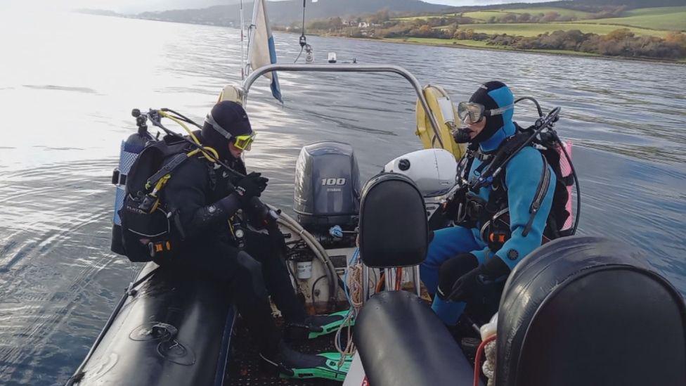 Two men in full diving gear, including oxygen tanks, sit on a rigid inflatable dingy ready to fall into the water