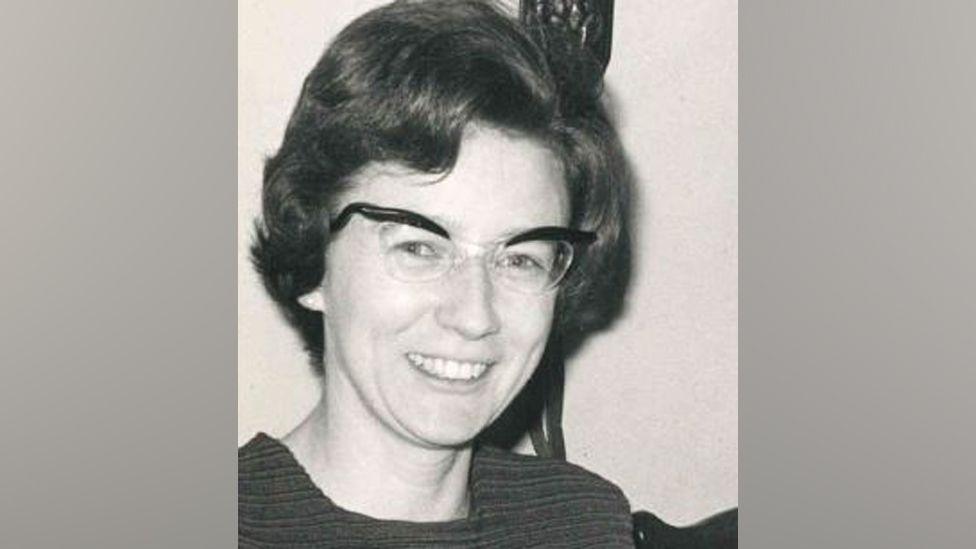 A black-and-white photograph of Myra Thompson. With dark hair and wearing glasses, she is smiling.