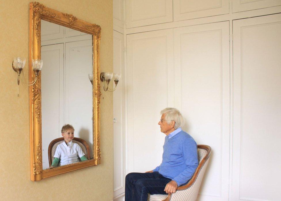 A man looks at a reflection of his younger self in the mirror