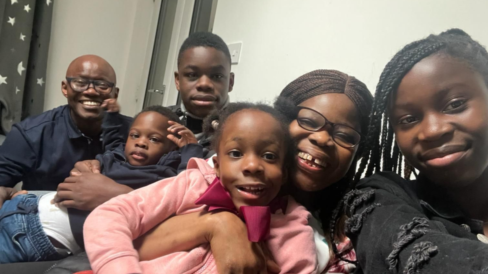 A man and woman sit down with their four children - two older and two toddlers. The woman is holding the younger girl while smiling and taking a selfie of all of them. 