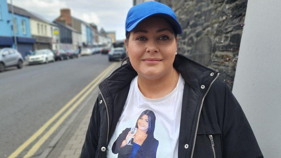 Karina Smith is standing on the side of Derry's Spencer Road and a number of cars and shops are visible in the background. She is wearing a black coat, a blue baseball cap and a white t-shit which has a picture of her niece Rebecca Browne printed on it