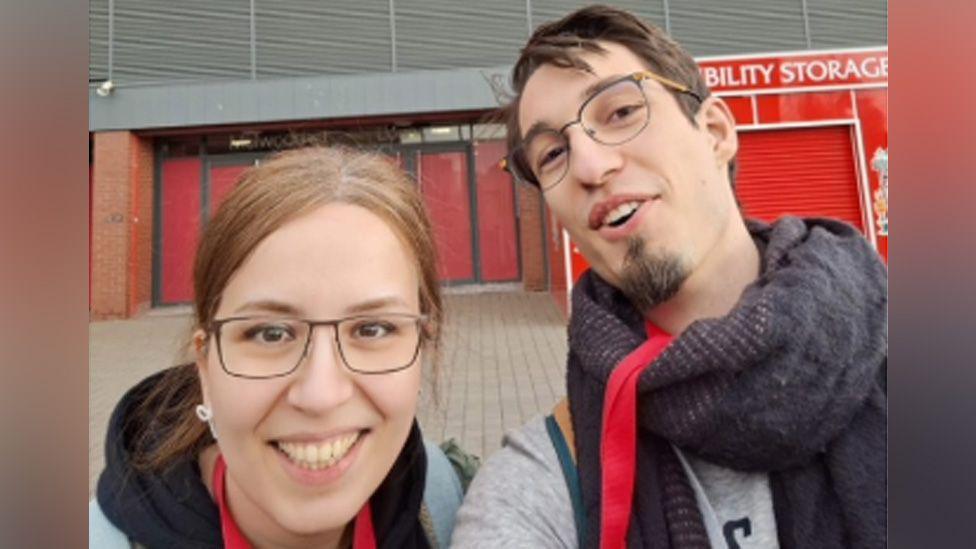 Dr Farnaz Kamranzad (L) has brown hair pulled and is wearing glasses and a black hooded top. Dr Antoine Septier (R) has short brown hair and is wearing glasses and a scarf. BOth are stood outside Anfield football stadium and smiling at the camera.