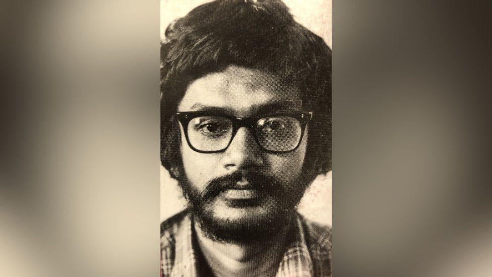 Viraj Mendis is staring directly at the camera in this black and white photograph. He is wearing black glasses and a chequered shirt. 