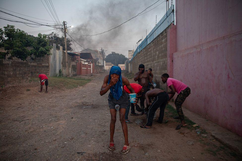 A man in blue shorts with a blue t-shirt over his head stoops and wipes his face with water. One man wearing red is bent over in the background to the left.  A group of five men do the same on the right by a wall painted pink. Smoke rises in the background by small buildings and a telegraph pole.