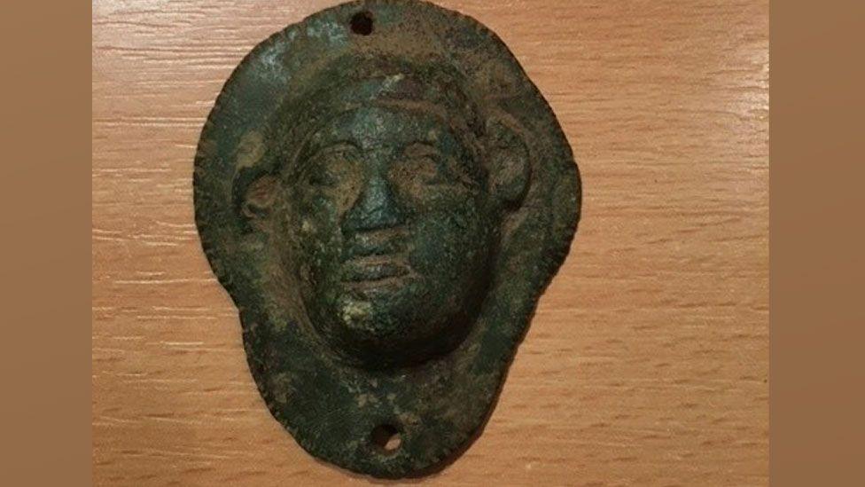 A Roman boss or harness decoration, decorated with a face with jug ears and with two attachment holes, one at the top and the other at the bottom