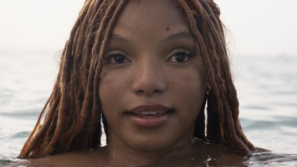 Halle Bailey as Ariel, in a close-up shot from The Little Mermaid. Her head is above water and the rest of her body is submerged. Her long, dreadlocked hair hangs down at each side of her head and disappears into the sea.