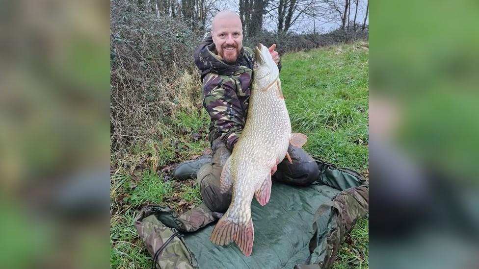 Lloyd Watson kneeling and holding up the pike