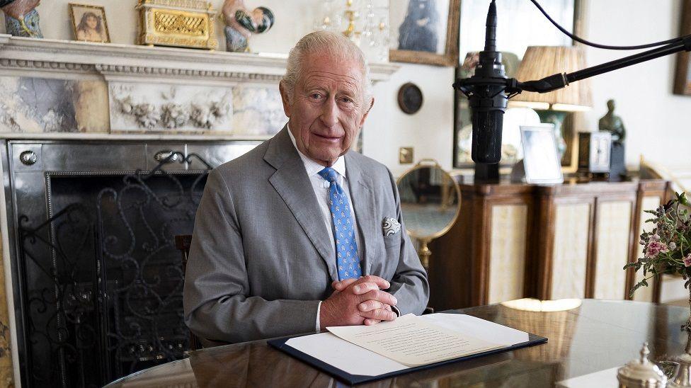 King Charles III wears a grey suit with a striking blue tie with his hands clasped at the foot of a piece of paper containing the words of his VJ Day message in the Morning Room of Clarence House, London. He is sitting in front of a marble-style fireplace. A microphone hovers near to this face.