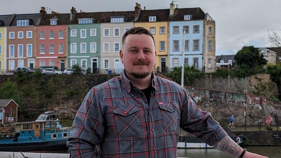 Alex Black standing on a boat with colourful buildings behind him