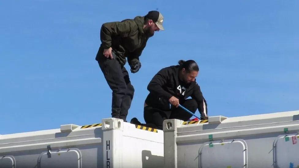 Two men using a crowbar on the lid of a white shipping container