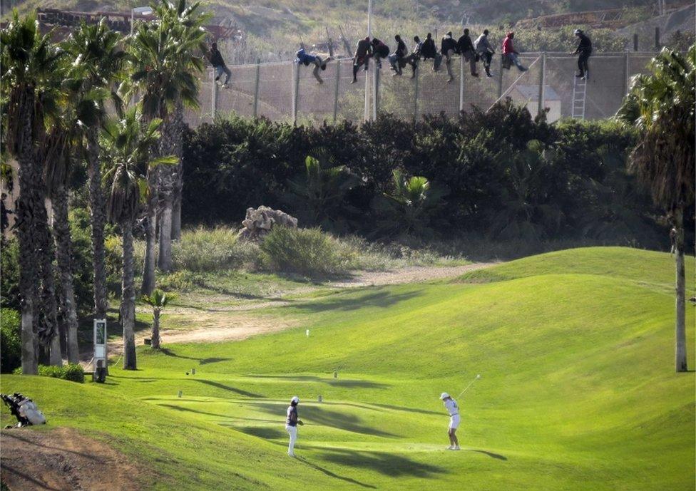 A golfer hits a tee shot as African migrants sit atop a border fence during an attempt to cross into Spanish territories between Morocco and Spain's north African enclave of Melilla