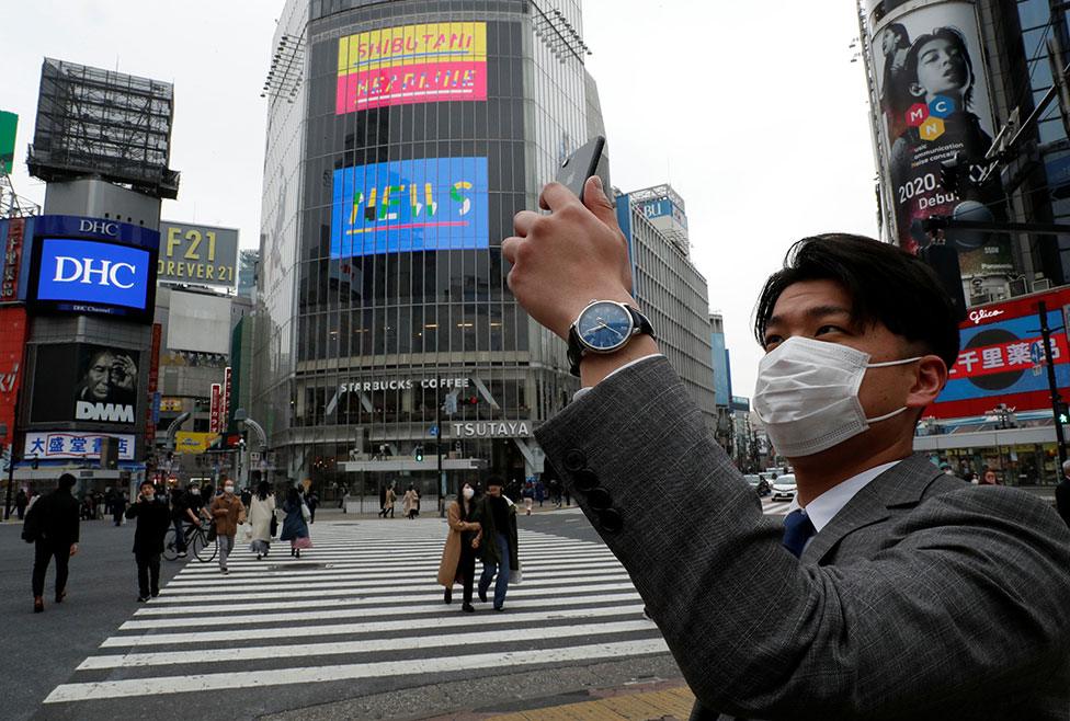 A man wearing a protective face mask takes a photo with his mobile phone at noon, at Shibuya Crossing in Tokyo, Japan