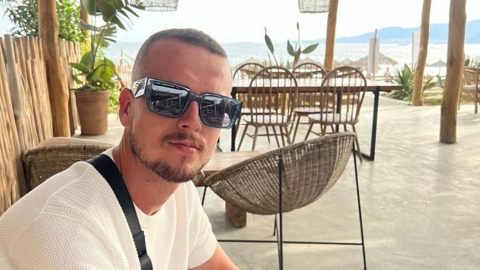 A young bearded man wearing a white t shirt and sunglasses. He is sat in what looks like a holiday resort with the sea and wooden furniture behind him.