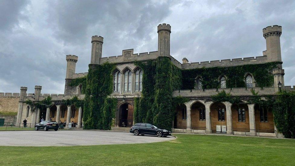 Exterior of Lincoln Crown Court a two-storey grey stone Victorian Gothic building with columns at the front, crenellated towers and vegetation growing along the walls. Two black cars are parked outside and a grass lawn sits in front of the building