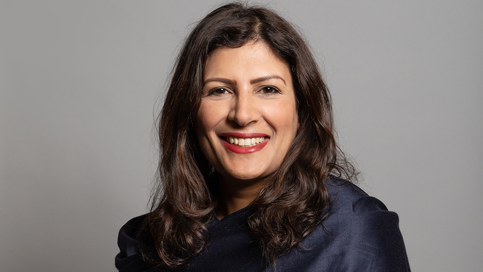 Head and shoulders shot of MP Preet Gill, wearing a dark top and standing against a grey background