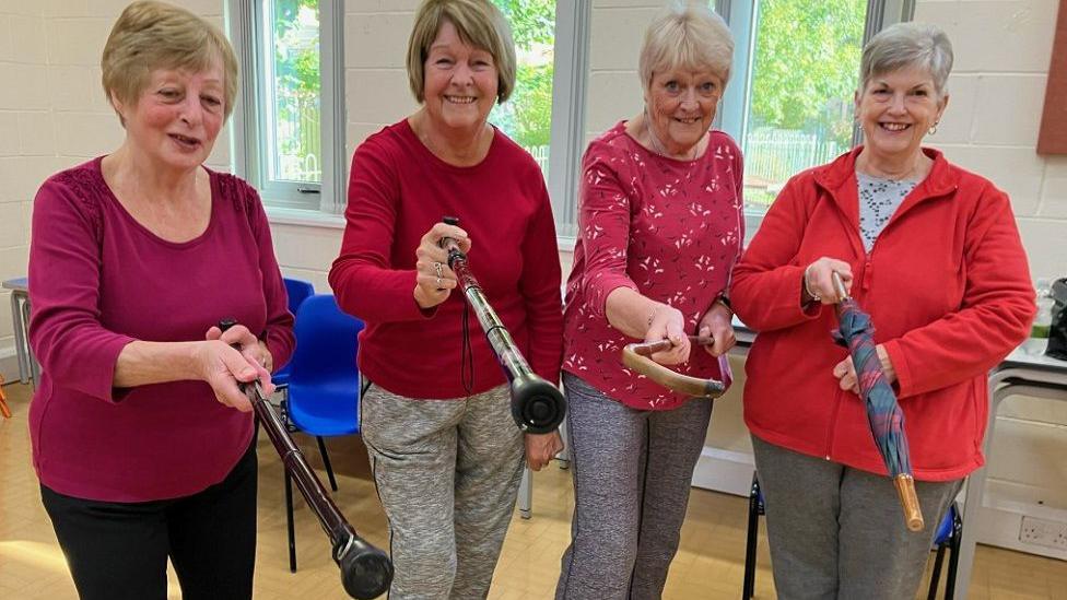 Carol, Sue, Ruth, and Joanne, all wearing pink/red tops with grey/black trousers and holding up walking sticks and umbrellas