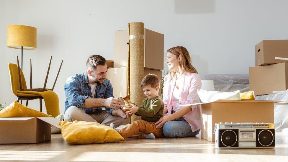 A couple with a young boy sitting on the floor in a room surrounded by boxes that they are unpacking. The man and the boy are both holding and looking at a bronze coloured jug