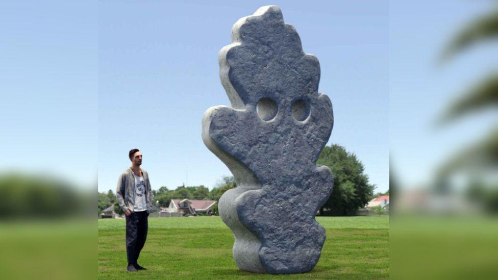 A computer generated image of what the installation will look like. It is a large, 13 foot tall sculpture made of stone. It is shaped irregularly like a leaf, with two large holes towards the top. A man is stood on the left, admiring the sculpture, which is positioned in a grassy field with homes and trees in the background.