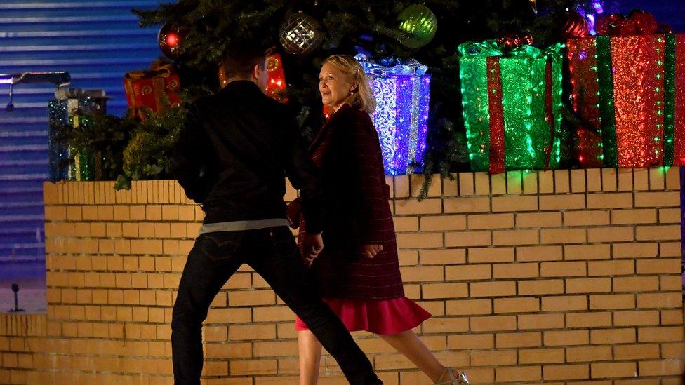 Gain and Stacey walk down a street at night and alongside a wall with Christmas decorations including presents