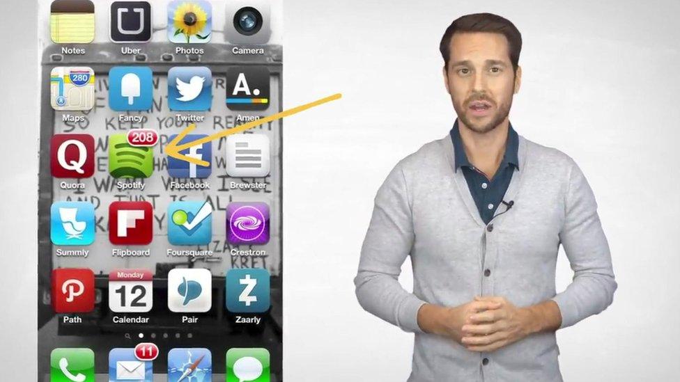 A man in a cardigan addresses the camera- he is depicted next to a graphic of a mobile phone with apps on display