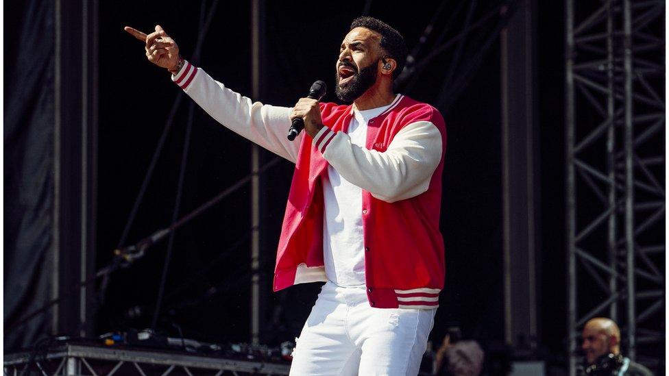 Craig David performing onstage wearing white trousers and a red jacket at Victorious Festival