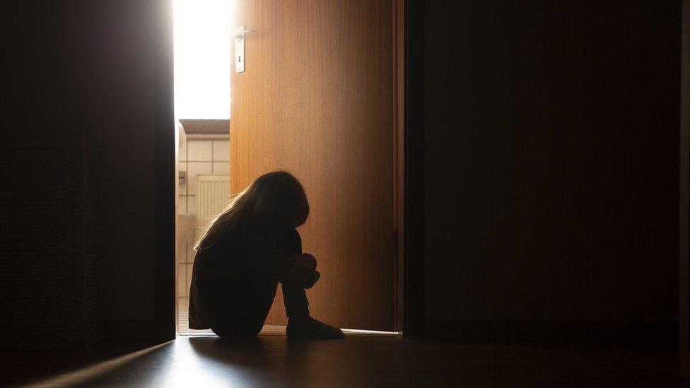 Despairing anonymous child sitting with head on knees in the dark frame of a doorway