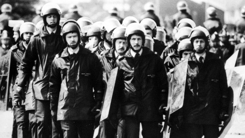 Police officers carrying riot shields walk towards a picket line