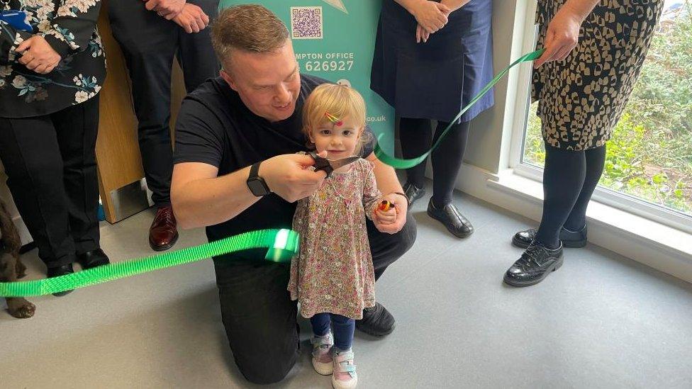 Chloe holds the scissors with her father kneeling beside her, having just cut a green ribbon as part of the hospital's unveiling