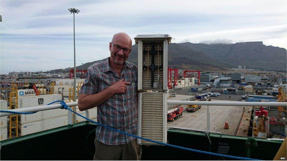 Peter Gibbs in front of a huge mountain, the sea in the background. He is onboard a ship and holding a large thermometer.