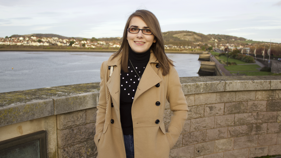 Elin standing in front of a wall with the sea behind her and houses and hills further away in the distance