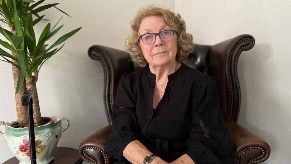 Elderly woman with curly brown hair and classis since in a leather armchair next to a pot plant