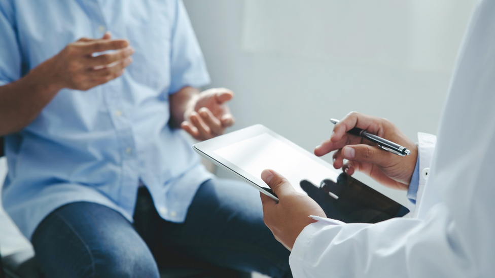 a doctor and a patient sitting down, the doctor is holding a tablet device