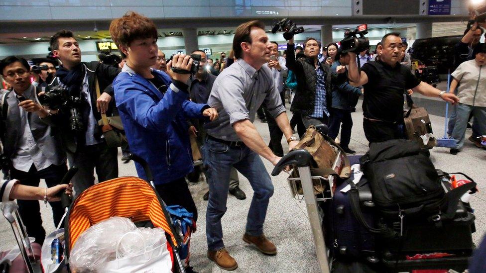 Rupert Wingfield-Hayes, pushing an airport trolley, surrounded by reporters and camera operators in the arrivals hall of Beijing International Airport