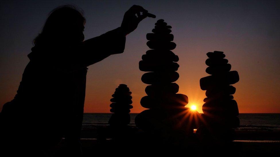 Woman adds pebble to sculpture as sunrises