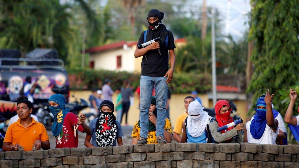 A young man, holding his homemade mortar, stands atop a makeshift wall, flanked by a dozen others. All wear face coverings.