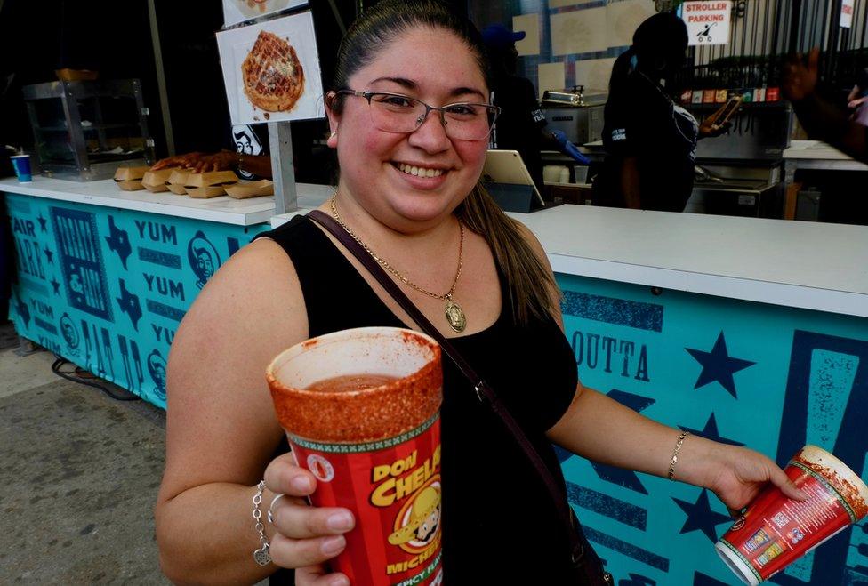 A woman holds a large paper cup filled with a red liquid. The rim has been dipped in spices