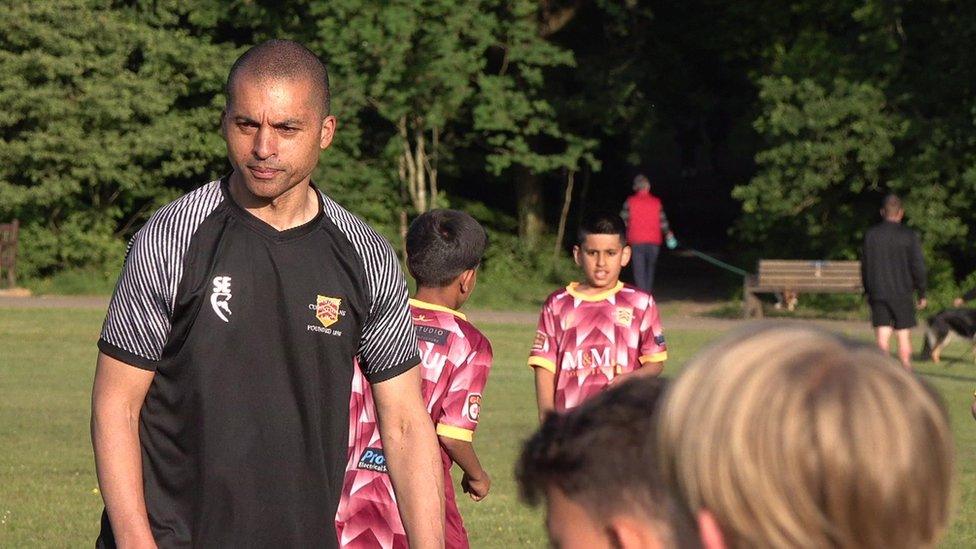 Spike is wearing a black and white football kit and is on a field, coaching some of the boys in the club.