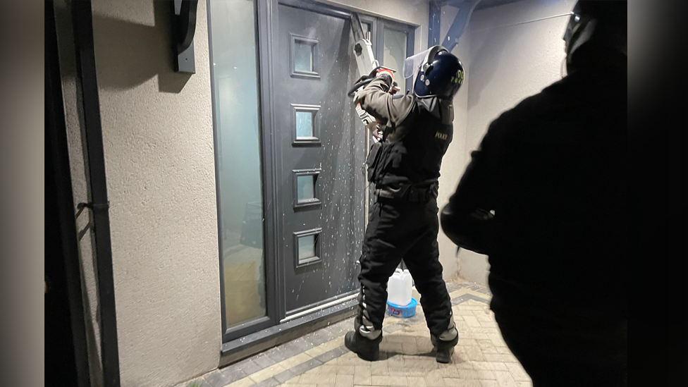 A police officer trying to break into a property with a chainsaw. There is another officer in the shot. The door is black with four small windows one of top of the other