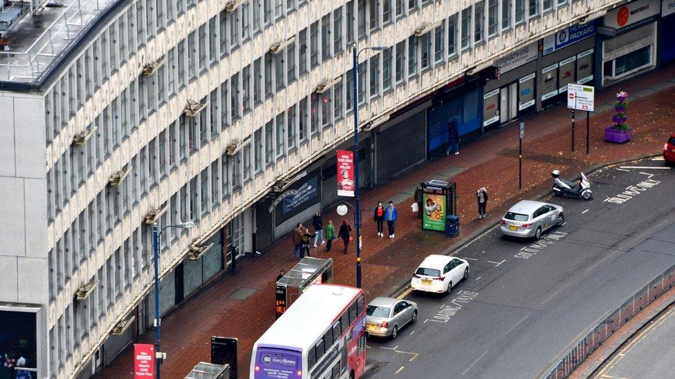 A four-storey grey building with tall thin windows on the top three layers, the bottom layer is blue shop fronts. A black and red brick pavement comes out from the shops and meets a tarmac road with cars and a bus on.