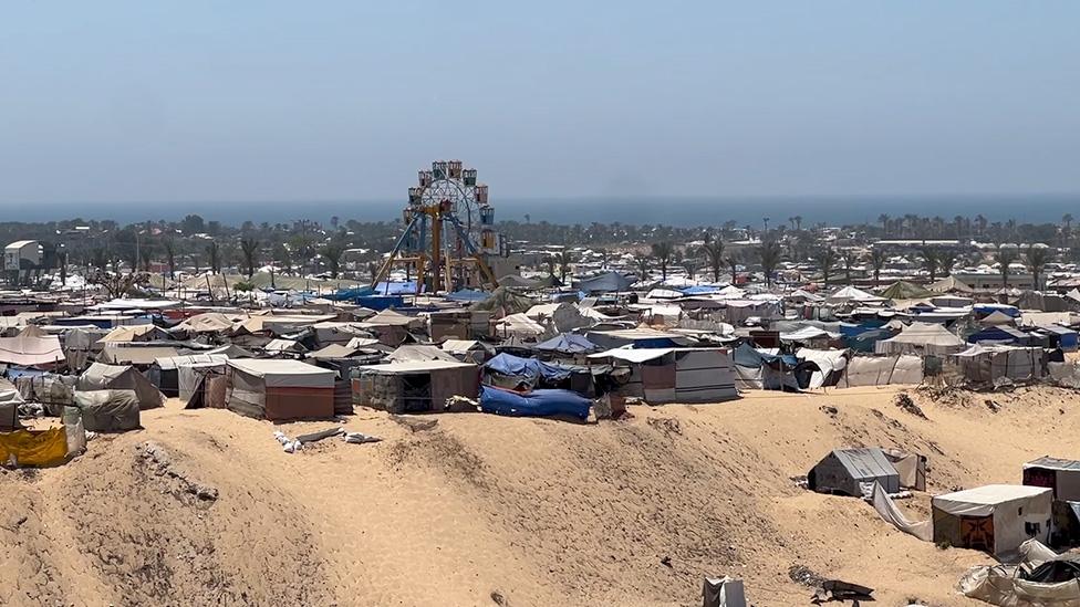 A wider shot showing the Ferris Wheel towering over a field full of white, brown and blue tents. Trees can be seen in the distance.