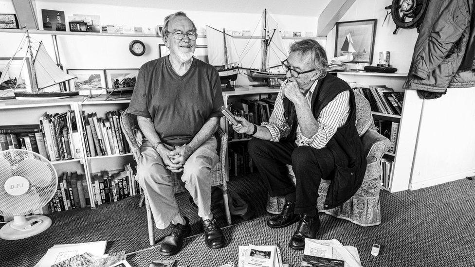 Black and white image of Tony James interviewing someone. They are sat side by side on chairs. Shelves crammed full of books can be seen behind them, as well as on the floor.