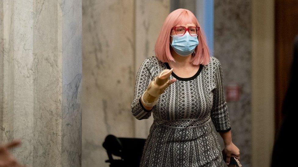 Kyrsten Sinema, wearing pink wig, red glasses and black-and-white dress, walks in a marble hallway.