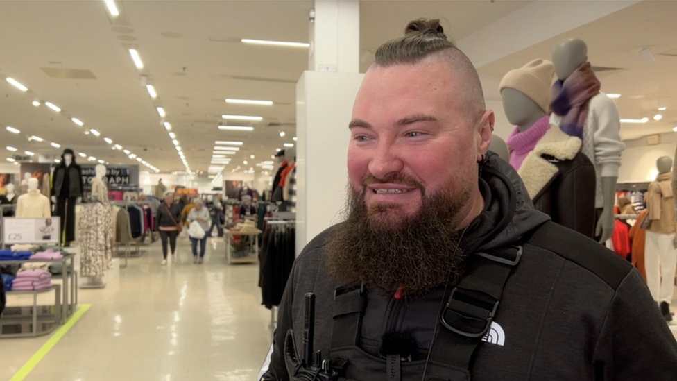 Sam is standing on the shop floor. He has his hair pulled back in a top knot bun and the sides are shaved. He has bushy black beard. He's known by locals as the Viking because of how tall he is! He is wearing a black hoodie and has a walkie talkie strapped to his chest.