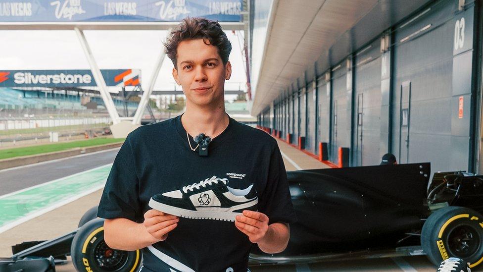 Alex Witty stands next to the Silverstone race track holding one of his new trainers with a Formula One car in the background