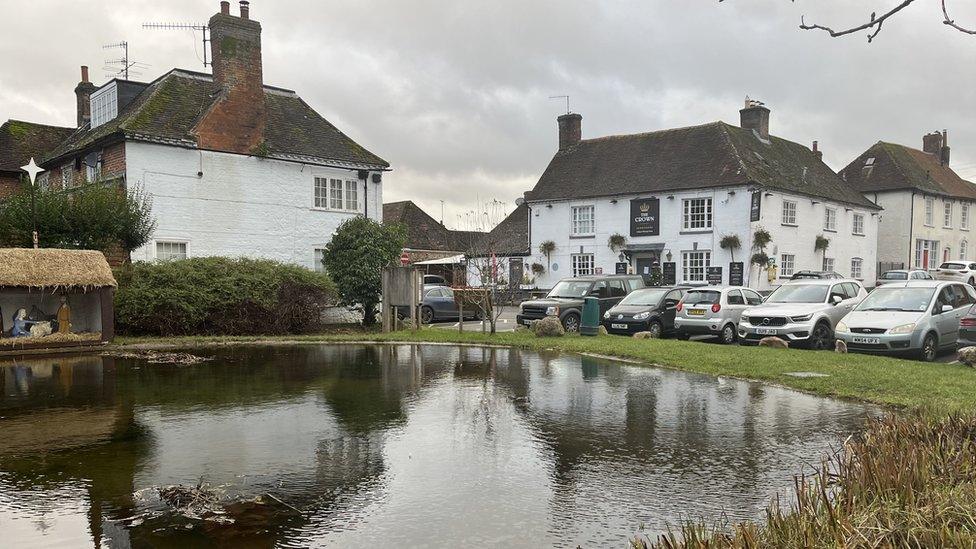 The centre of Aldbourne with a pond, old whitewashed pub and houses and parked cars