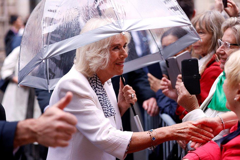 Queen Camilla shakes hands with people as she walks in a street